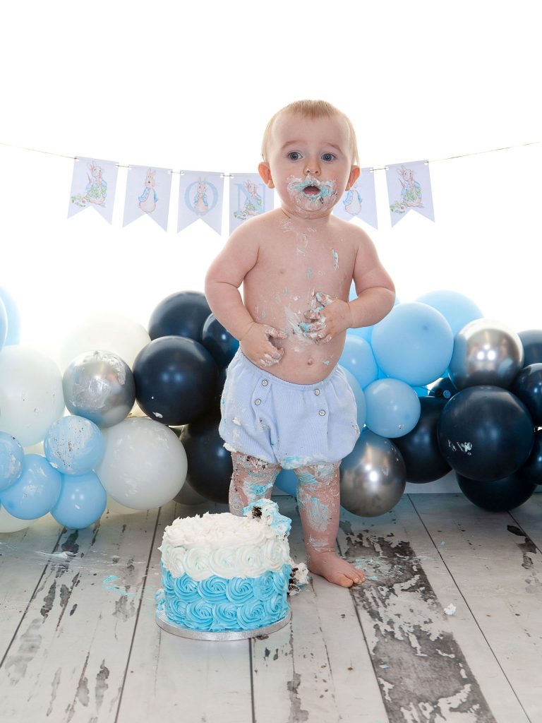 Little baby boy stood up with cake all over his legs, tummy, hands and face